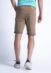 Shorts de mezclilla color arena con puños doblados, que cuentan con dos bolsillos traseros y una etiqueta en la cinturilla. Llevados con zapatillas de color claro.