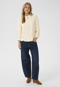 Cremefarbene Bluse mit strukturiertem, kariertem Muster, Knopf-Front und Rüschencollier; kombiniert mit dunkelblauen weit geschnittenen Jeans und braunen Stiefeletten.