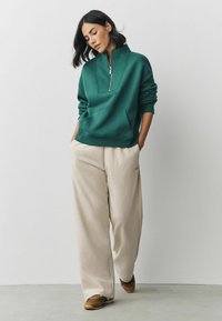 Grøn zip-up sweatshirt med en kengurulomme, parret med cremede brede bukser og brune sneakers. Enkel, afslappet urban stil.