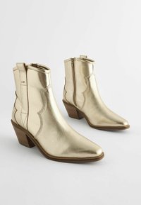 Botas de tobillo metálicas doradas con puntas afiladas y un diseño con volantes. Cuentan con cremalleras laterales y un tacón en bloque texturizado.