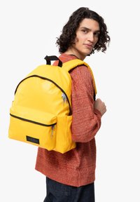 Mochila amarilla con una textura suave, bolsillo frontal prominente, detalles de cremallera y ribete negro. Llevada por una persona con un peinado rizado.