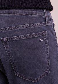 Los pantalones vaqueros de mezclilla gris oscuro presentan un bolsillo trasero con un sutil detalle de costura y un pequeño logo. La textura parece suave con un corte clásico.