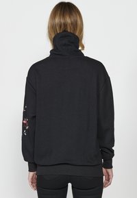 Sweatshirt noir avec un col montant et une texture en chevrons ; présente des broderies florales sur la manche gauche ; coupe loose et décontractée.