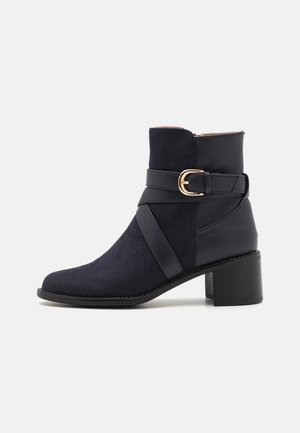 Black ankle boot with 5 cm block heel, suede front, leather back, and decorative strap with gold buckle on white background.