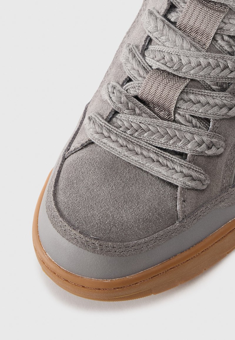 Gray suede sneaker featuring braided gray laces, a smooth gray leather trim, and a brown rubber sole. Close-up view of upper section.