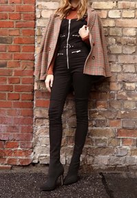 Blazer marrón a cuadros, suéter de cuello alto naranja, overoles negros con cremalleras plateadas y cinturón en la cintura, pantalones ajustados negros y botas altas de tacón negras.