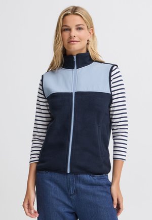 Femme portant un gilet sans manches bleu marine et bleu clair zippé sur une chemise blanche à manches longues avec des rayures horizontales noires et un jean bleu.