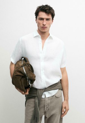 Junger Mann trägt weißes kurzärmeliges Hemd, beige Hose, grauen Pullover um die Taille gebunden, hält eine braune Wildledertasche mit Reißverschluss.