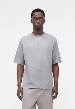 Ung mand iført en ensfarvet lysegrå kortærmet T-shirt og lysegrå bukser, stående mod en hvid baggrund.