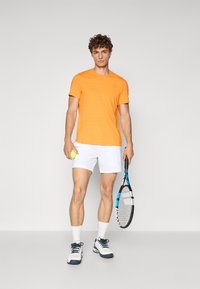 Oranž T-särk lühikeste varrukatega, millel on peened horisontaalsed triibud, koos valgete lühikeses pükste komplektiga. Mudel hoiab tennisereketit ja tennisepalli.