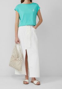 Kvinna som bär en turkos kortärmad topp och en vit maxikjol med sprund fram, håller en beige nätväska och bär vita slidesandaler.