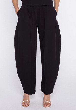 Kobieta w luźnych czarnych spodniach z bocznymi kieszeniami i złotych sandałach na paski z odkrytymi palcami, stojąca na tle jednolitego białego tła.