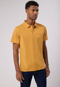 Polo à manches courtes de couleur jaune vif, fabriqué dans un matériau doux. Présente une patte de boutonnage à trois boutons et un petit logo sur la poitrine.