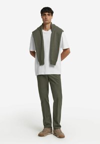 Weißes T-Shirt, olivgrüner Strickpullover über die Schultern drapiert, olivgrüne Hose, hellbraune Sneakers. Einfaches und lässiges Outfit.