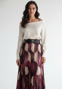 Kremowa bluzka z odkrytymi ramionami w połączeniu z plisowaną bordową spódnicą ozdobioną okrągłymi wzorami w kolorze białym i różowym. Czarny pasek z okrągłą klamrą.