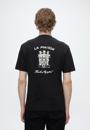 Person with curly hair wearing black T-shirt featuring white sketch of a building and text "LA MAISON Karl Lagerfeld" on the back.
