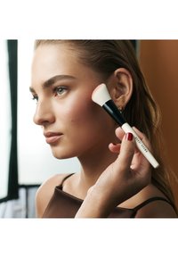 En makeupborste applicerar rouge på en modells kind. Borsten har ett svart handtag och vita borst. Modellen har slät hud och en brun topp.