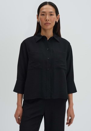 Frau trägt ein locker sitzendes schwarzes Hemd mit weiten Manschetten und zwei aufgesetzten Brusttaschen, steht vor einem schlichten hellen Hintergrund.