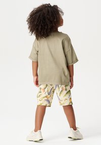 T-shirt vert olive clair à manches courtes, associé à un short crème orné de motifs colorés de fruits et de feuilles, et des baskets blanches.