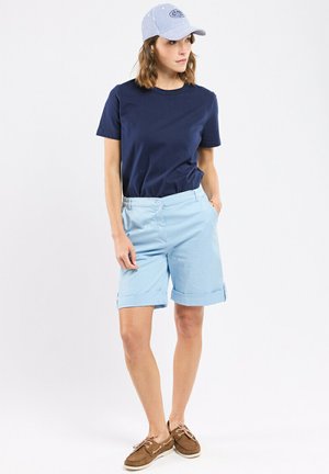 Femme portant un t-shirt bleu marine, un short bleu clair retroussé, une casquette rayée et des chaussures bateau marron, debout avec une main dans la poche.
