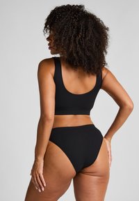 Donna con capelli ricci che indossa un bikini a coste nero, vista di spalle, in piedi contro uno sfondo grigio uniforme.
