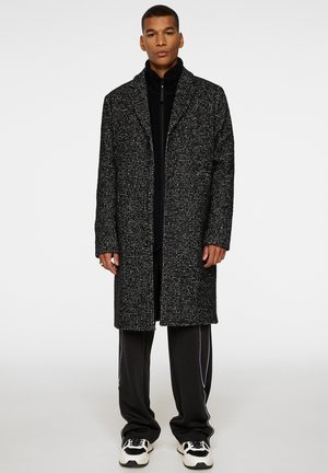 Svart och vit strukturerad kappa med lång silhuett, notchade slag och knäppning, bärs över en svart zip-up topp och mörka byxor.