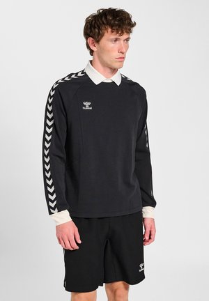 Mężczyzna ubrany w czarny strój sportowy z białymi paskami w kształcie jodełki na rękawach i krótkich spodenkach, stojący na jednolitym jasnym tle.
