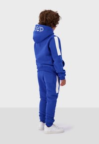 Blue hoodie and pants set made of soft fabric, featuring white side stripes. Hood has logo detail. Paired with white sneakers.