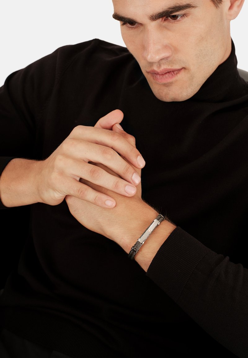 Bracciale intrecciato argento e nero con una chiusura metallica rettangolare elegante, indossato su un polso con le mani unite. Visibile un semplice maglione nero.