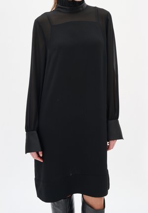 Kobieta w czarnej sukience z długimi rękawami, przezroczystymi górnymi rękawami i wysokim kołnierzem, w połączeniu z czarnymi kozakami za kolano.