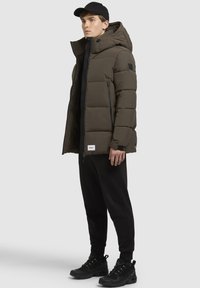 Puffer jacket in dark olive green, with a high collar, horizontal quilting, zip pockets, and black accents. Paired with black joggers and sneakers.