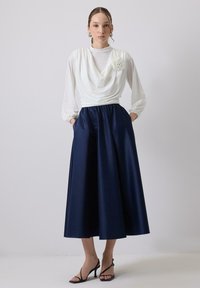 Top bianco drappeggiato con dettaglio di rosa, abbinato a una gonna fluente in satin blu navy. Presenta maniche lunghe e tasche laterali. Tacchi neri con cinturini.