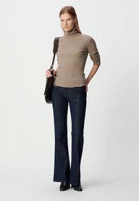 Beige geribbelde coltrui, slim fit. Donkerblauwe uitlopende jeans. Zwarte enkellaarsjes. Model houdt een zwarte tas vast. Simpele, neutrale achtergrond.