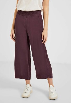 Personne portant un pantalon ample, raccourci, de couleur violet foncé avec une taille élastique et des baskets blanches sur un fond uni.