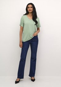 Tricou V verde deschis din material moale, asociat cu blugi evazați de un albastru închis. Modelul stă cu o mână pe șold, purtând pantofi cu toc stiletto negri.