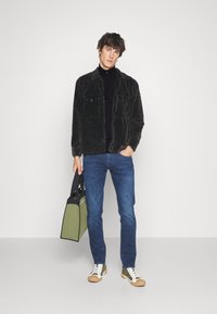 Schwarze Cordjacke mit Brusttaschen, kombiniert mit einem schwarzen Rollkragenpullover und blauen Jeans. Die Schuhe sind weiß mit grünen Akzenten. Hält eine grüne Tasche.