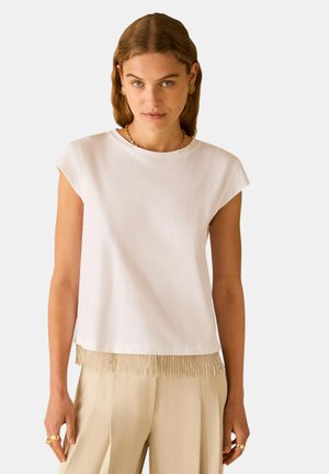 Donna che indossa un top bianco senza maniche con orlo a frange e pantaloni beige a gamba larga, accessoriata con gioielli dorati.