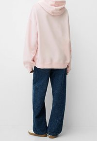 Růžová mikina s volným střihem a kapucí na stažení, spárovaná s texturovanými modrými kalhotami s širokými nohavicemi a světlými teniskami.