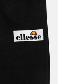 Melnas audums ar baltu taisnstūra plāksteri, uz kura ir "ellesse" logo melnā krāsā un stilizēts puslocals oranžā un dzeltenā krāsā.