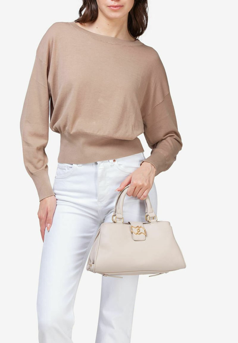Beige struktureret håndtaske med guldfinish, to overhead-håndtag og lynlåslukning. Modellen bærer en lys sweater og hvide bukser. Glat læderfinish.