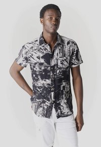 Jeune homme debout, main dans la poche, portant une chemise à manches courtes noire et blanche à motifs et un short blanc déchiré, sur fond uni.