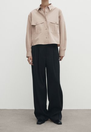 Person wearing beige cropped button-up shirt with large chest pockets and wide-leg black trousers standing on gray floor against white wall.