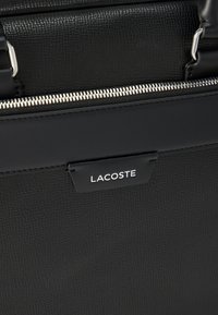 Gros plan sur un sac en cuir noir texturé avec une fermeture éclair argentée visible et une étiquette noire affichant le nom de la marque Lacoste en lettres blanches.
