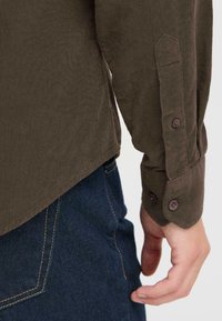 Camisa de corduroy verde oliva oscuro con puños abotonados, con canalé vertical y un dobladillo ligeramente curvado, combinada con unos vaqueros de denim azul.