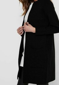 Žena v dlouhém černém kardiganu přes bílou košili, držící přední okraje kardiganu oběma rukama.