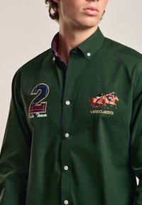 Camisa de botones de color verde oscuro con un cuello de cuadros. Presenta un número "2" bordado y "Polo Team" en un lado, con un motivo de caballo y el texto "valecuatro".