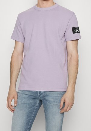 Lila Baumwoll-T-Shirt mit strukturierter Oberfläche, Rundhalsausschnitt, kurzen Ärmeln und einem schwarzen Logo-Patch am linken Ärmel. Kombiniert mit hellblauen Jeans.