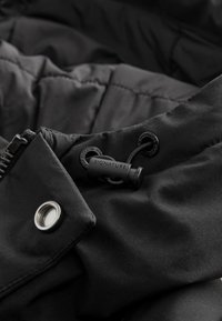 Czarna tkanina kurtki ocieplanej z pikowaną teksturą; posiada zamek błyskawiczny i sznurek ze szczegółami metalowymi z logo marki. Akcent z metalowym zatrzaskiem.