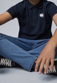 Granatowa koszulka polo z trzema guzikami i naszywką z logo, zestawiona z niebieskimi dżinsami i czarnymi trampkami z białymi sznurowadłami i czerwonymi akcentami.