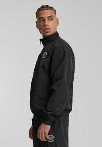 Svart zip-up jacka med hög krage, med elastiska ärmslut och en rund logotypapplikation på bröstet. Tillverkad av lätt, strukturerat material.